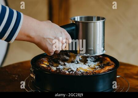 El primer plano de una mano femenina rocía azúcar en polvo sobre una torta sobre una mesa marrón de madera Foto de stock