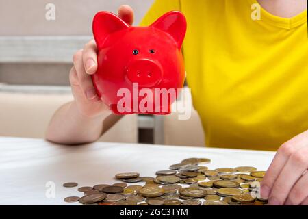 Las manos femeninas sacuden un banco piggy para comprobar el contenido, varias monedas yacen en una mesa en casa. Foto de stock