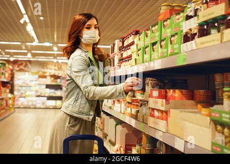 Una mujer bonita escogiendo comestibles en el supermercado. Las consumidoras femeninas compran bienes en un centro comercial, caminando entre estanterías. Chica con máscara de compras de alimentos