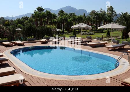Vista a la piscina vacía, tumbonas y sombrillas en un jardín con palmeras. Vacaciones en complejo tropical sobre fondo de montañas
