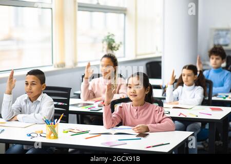 Un grupo diverso de pequeños escolares levantando las manos en el aula Foto de stock