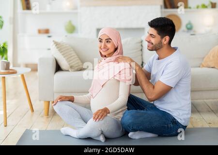 Amoroso marido árabe masajeando los hombros de su mujer embarazada en casa, sentados juntos en el suelo. Hombre tierno dando un masaje a su encantadora esposa para revivir