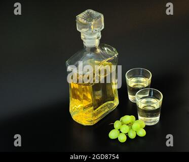 Grappa, un alcohol fuerte a base de uvas blancas, un decantador de vidrio, dos vasos y un manojo de bayas sobre fondo negro. Primer plano. Foto de stock