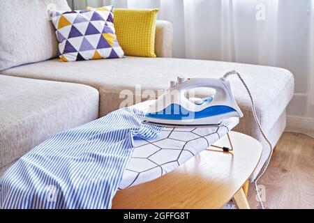 Toallas lavadas en una tabla de planchar y una plancha en una pequeña sala  de estar con sofá, tiempo de rutina del hogar