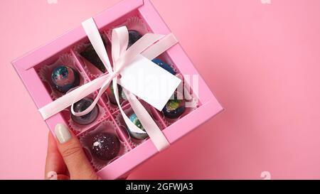 Dulces de chocolate. La mano sostiene una caja de regalo rosa con chocolates hechos a mano sobre un fondo rosa. Fondo festivo. Etiqueta blanca. Burlándose Foto de stock