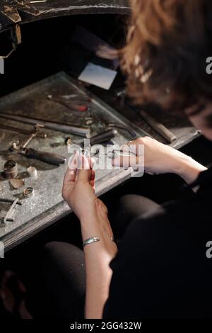 Vista sobre el hombro del joyero hembra del banco usando pulidor de mano mientras que realiza la reparación de la joyería en taller Foto de stock