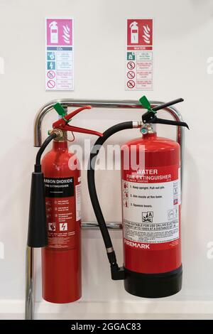 Un par de extintores rojos - agua y dióxido de carbono - en un soporte de apoyo en una oficina con avisos / señales de seguridad, Reino Unido Foto de stock