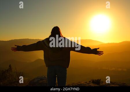 Vista posterior silueta con retroiluminación de una mujer estirando los brazos al atardecer en invierno