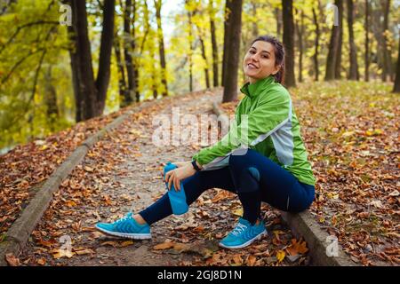 Corredor descansando después del entrenamiento en el parque de otoño. Mujer feliz sosteniendo una botella de agua sentada en el camino. Estilo de vida deportivo activo y saludable. Entrenamiento de otoño