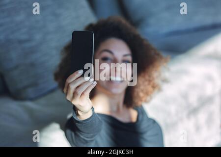 Feliz mujer joven tomando un selfie en su teléfono móvil como ella relaja en el suelo apoyado en un ofa con el foco al smartphone Foto de stock