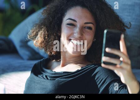 Divertida mujer joven riendo de su teléfono móvil mientras se relaja en el suelo con un rayo de sol cálido en la sala de estar de casa Foto de stock
