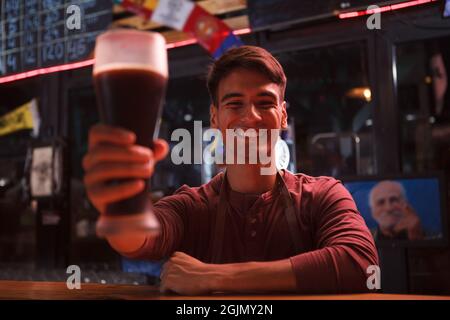 Feliz joven barman sonriendo, sosteniendo un vaso de cerveza en la cámara Foto de stock