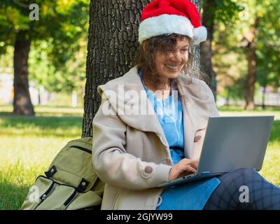 Foto recortada de una mujer de mediana edad en sombrero de Santa con videollamada en línea. Ella sentada en la hierba bajo el árbol en el parque de la ciudad con ordenador portátil. Moderno t