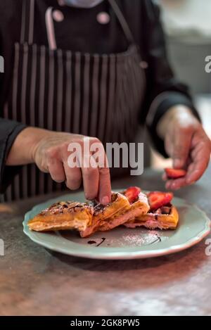 Cocina masculina anónima poniendo rodajas de fresa madura en waffles vieneses con salsa de chocolate y azúcar en polvo en el plato en la cocina