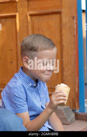 Alegre niño contento en una camisa azul come helado en el porche de una casa en un pueblo en un día soleado de verano. Enfoque selectivo. Vertical Foto de stock