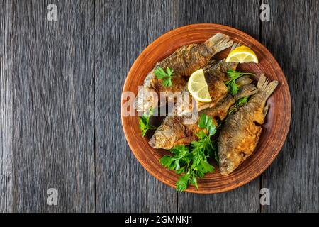 carpas de crucería fritas con corteza dorada crujiente en un plato rústico de arcilla con perejil y limón, capa plana Foto de stock