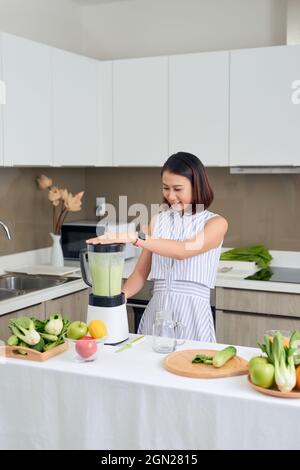 Vegan sonriente mujer asiática haciendo un batido con fruta y verdura en la cocina