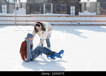 longitud completa de hombre sonriente cayendo cerca de la mujer mientras patinan en la pista de hielo afuera Foto de stock