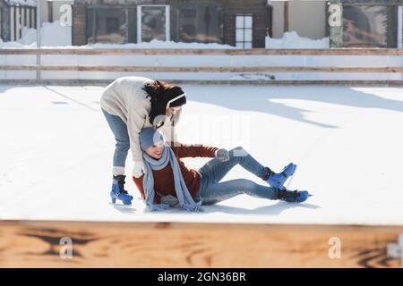 longitud completa de hombre feliz cayendo cerca de la mujer mientras patinan en pista de hielo fuera Foto de stock