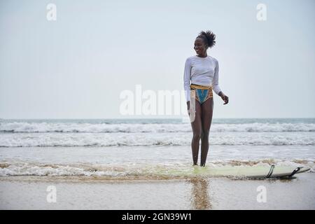 Alegre joven deportista étnico en bikini de pie en la costa del mar contra la tabla de surf mientras mira bajo el cielo claro Foto de stock