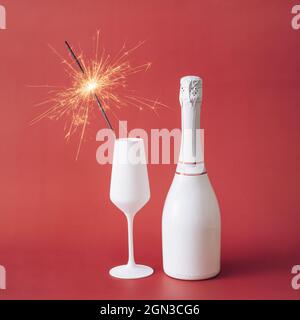 Primer plano de un sparkler en una flauta blanca junto a una botella de champán sin etiquetar contra fondo rojo para espacio de copia como símbolo de alegría en el celebrat Foto de stock