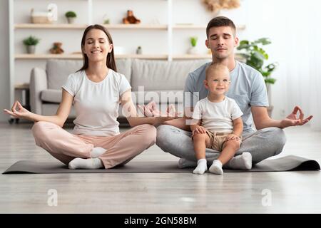Padres jóvenes relajados y bebés pequeños meditando juntos en el hogar Foto de stock