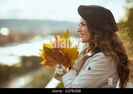 Hola septiembre. Sonriente mujer de mediana edad moderna en gabardina beige y beret negro con hojas amarillas de otoño al aire libre en la ciudad en otoño.
