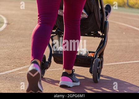Madre con leggings rosa y zapatos empujando a su hijo en un cochecito a lo largo del camino por la mañana Foto de stock