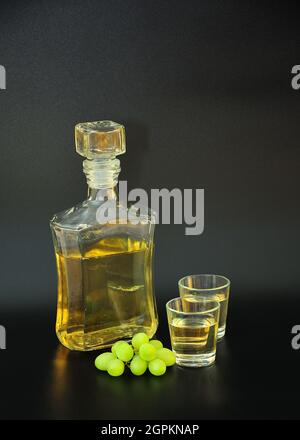 Grappa, una botella de vidrio y dos vasos de alcohol fuerte sobre fondo negro, junto a un manojo de uvas blancas maduras. Primer plano. Foto de stock