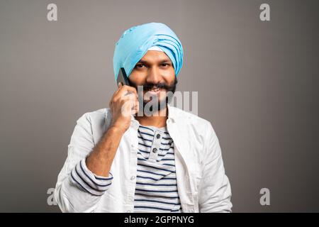 Feliz sonriente indio Sikh hombre ocupado hablando en el teléfono móvil en el fondo del estudio por mirar la cámara - concepto de comunicación, red y positivo Foto de stock