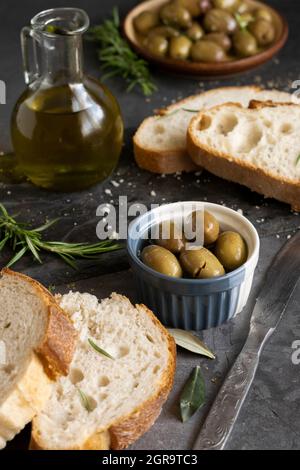 Pan italiano en rebanadas de ciabatta en tabla de cortar con hierbas, aceite virgen extra y aceitunas marinadas