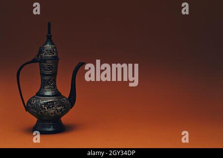 Cántaro tradicional islámico oriental grabado hecho a mano sobre fondo naranja oscuro. Elegante jarra arábiga de metal alto, espacio para copias.