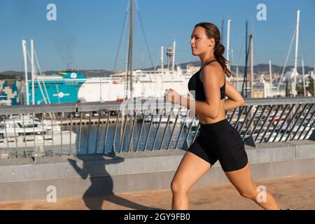 Deportista centrado en la ropa deportiva que corre sobre el terraplén durante el entrenamiento cardiovascular en la mañana sin nubes en el puerto. Foto de stock