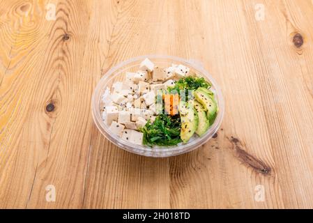 Cuenco vegano con cubos de tofu, ensalada de algas wakame, aguacate, semillas de sésamo y amapola con arroz blanco y huevas de masago sobre mesa de madera Foto de stock
