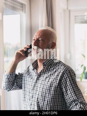 Anciano emocionado en camisa con pelo blanco y barba hablando en el teléfono junto a la ventana en casa Foto de stock