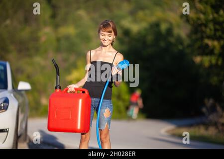 Feliz niña de pie al lado de un coche eléctrico. Sujetando el cable de carga y el contenedor de gascolina. Foto de stock