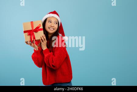 Feliz mujer asiática en Santa sombrero sosteniendo el presente y sacudiendo la caja de golpe, mirando a un lado el espacio libre sobre fondo azul Foto de stock