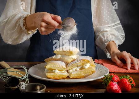azúcar glaseado en polvo rociado de un tamiz en la mano de la mujer, cocinando hojaldre dulce con crema Foto de stock