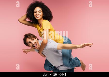 feliz mujer latina piggybacking en asombrado novio imitando el avión con las manos estiradas aisladas en rosa Foto de stock