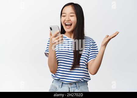 Imagen de una chica castaña asiática mirando con cara excitado y sorprendida al smartphone, reaccionando asombrada ante la notificación del teléfono móvil, de pie sobre blanco Foto de stock