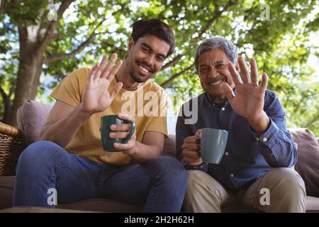 Feliz hijo adulto biracial y padre mayor sosteniendo tazas de café y haciendo videollamada con computadora portátil