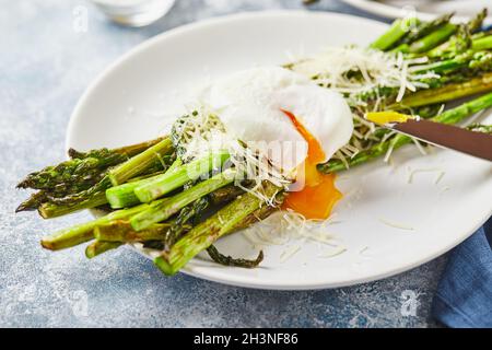Espárragos verdes con huevo escalfado y parmesano, desayuno vegetariano servido en dos platos blancos sobre fondo claro. Foto de stock