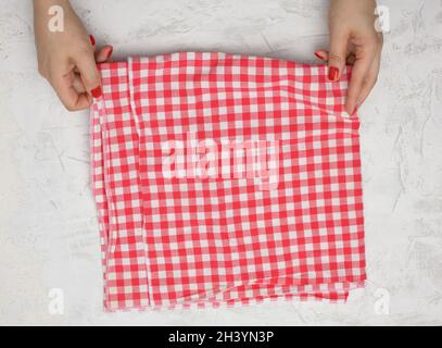 Dos manos femeninas sostienen una servilleta de cocina textil a cuadros de color blanco y rojo arrugada sobre una mesa blanca Foto de stock