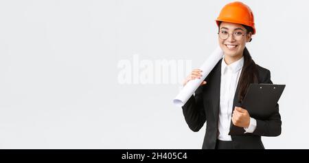 Arquitecto asiático exitoso en traje de negocios y casco con blueprints y portapapeles con notas, inspector mirando