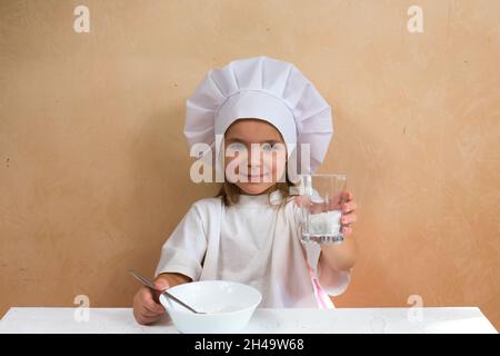 Una chica sonriente y alegre vestida como cocinera muestra un vaso de harina para amasar la masa. Foto de stock