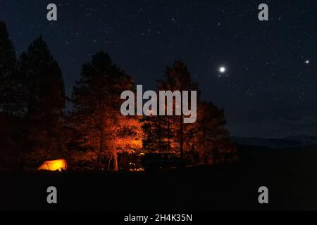 Vista nocturna panorámica del campamento turístico con carpa naranja iluminada, alerces amarillos, hoguera y coche en la cima de las montañas contra el cielo estrellado Foto de stock