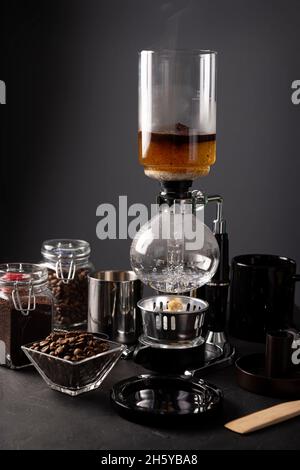 La cafetera de vacío también conocida como cafetera vac, sifón o sifón y granos de café tostados sobre una mesa rústica de piedra negra. Foto de stock