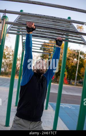 Hombre de pelo gris fuerte con relojes reorganiza sus manos en la barra horizontal de mono al aire libre. Estructura de escalada para actividades deportivas al aire libre. Adulto Foto de stock