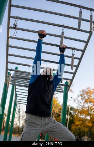 Hombre de pelo gris fuerte con relojes reorganiza sus manos en la barra horizontal de mono al aire libre. Estructura de escalada para actividades deportivas al aire libre. Adulto Foto de stock