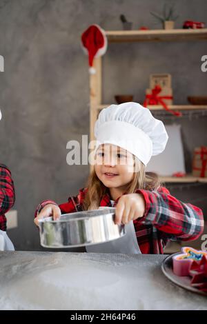 la niña sifts harina para la masa a través de un tamiz en la mesa. El niño cocina en la ropa del chef. Niños pasteles caseros en la cocina. Foto de stock